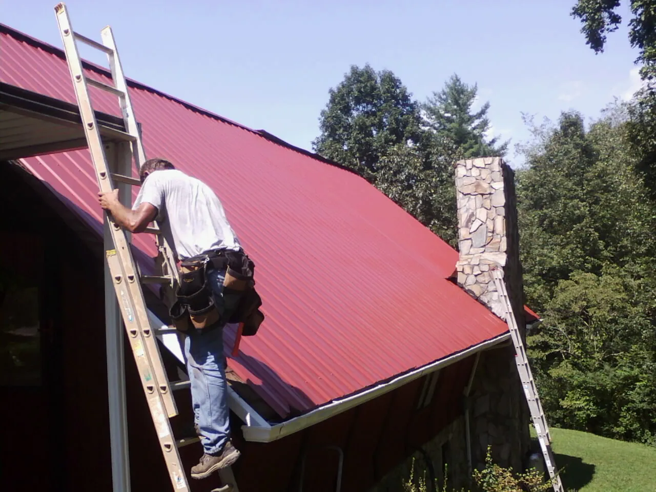 Roofer on a ladder beside a red metal roof and stone chimney — Soffit & Fascia Repair by Roofing Brothers in Arden